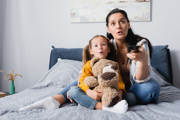 excited mother and daughter watching tv while sitting on bed with teddy bear