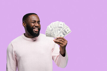 Satisfied african american man holds bunch of money in his hand enjoying of winning or salary, standing on isolated background and winking an eye at the camera