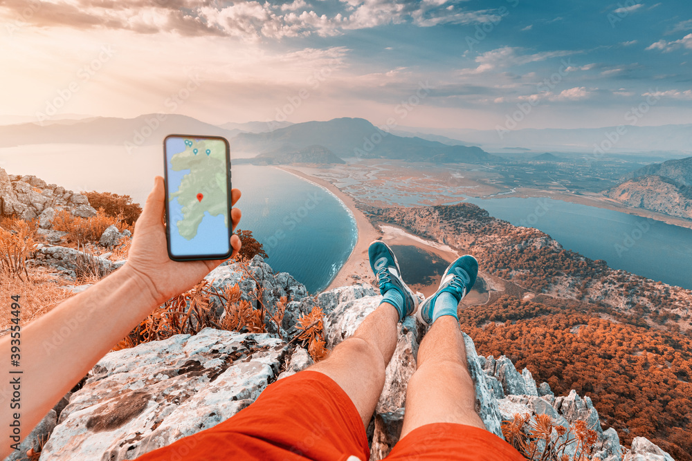 Young and sportive man on top of a mountain with a stunning view of the Iztuzu beach in Turkey and the sea coast looks into a smartphone with a map app for navigation - obrazy, fototapety, plakaty 