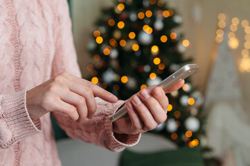 The girl holds her finger in front of the smartphone. against the background of a Christmas tree with festive decor.