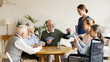 © Comeback Images - Young nurse bringing vitamin pills for senior man playing cards with aged friends sitting at table in common room of nursing home