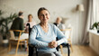 © Comeback Images - Portrait of disabled senior woman in wheelchair looking at camera and smiling happily in nursing home; other aged patients in background