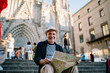 © BullRun - Portrait of happy male tourist with location map in hands smiling at camera during solo vacations in Spain, cheerful man in straw hat posing in historic center spending daytime for exploring town
