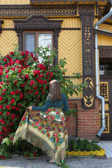 Wall Mural - Old ornamental windows, carved frames. Vintage wooden rural house in Suzdal town, Russia. Decorative red climber roses, bush: season flowers in summer garden. Russian girl in folk Pavlovo Posad shawl