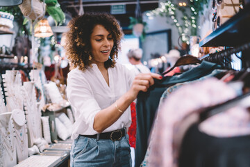 Attractive trendy dressed woman choosing clothes in fashion store concentrated on decision for shopping, stylish curly female searching wear in showroom of talented designer creating trendy look