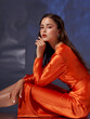 © Dmitry Tsvetkov - Studio portrait. Elegant woman sitting and posing in orange silk dress against blue metaallic background. High fashion female model with make-up and wet hairstyle
