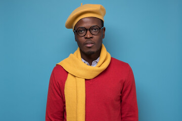 Wall Mural - Serious African man in glasses, scarf and yellow hat looking at camera. Studio shot on blue wall.