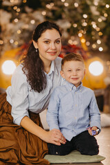 Happy mother with son sit on floor against background of glowing headlights and Christmas decorations.