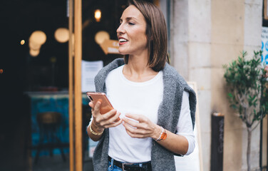 Wall Mural - Pretty female blogger dressed in trendy wear holding modern cellphone device and looking away, charming millennial girl 30 years old using mobile device enjoying weekend and leisure time in city