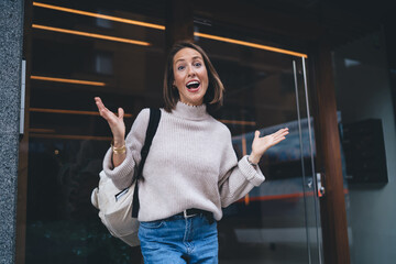 Wall Mural - Half length portrait of astonished female backpacker raising hands showing own emotion while waiting friend in touristic city, Caucasian woman dressed in casual apparel surprised with shock news