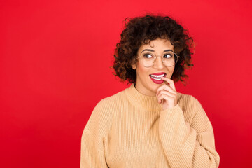 Sticker - Young beautiful Arab woman wearing beige sweater against red background with thoughtful expression, looks to the camera, keeps hand near face, bitting a finger thinks about something pleasant.