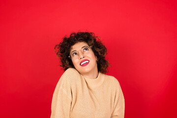 Wall Mural - Portrait of mysterious Young beautiful Arab woman wearing beige sweater against red background looking up with enigmatic smile. Advertisement concept.