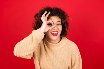 Sticker - Young beautiful Arab woman wearing beige sweater against red background with happy face smiling doing ok sign with hand on eye looking through finger.