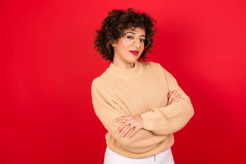 Wall Mural - Waist up shot of  self confident Young beautiful Arab woman wearing beige sweater against red background has broad smile, crosses arms, happy to meet with colleagues.