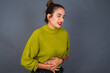 © Roquillo - Young beautiful hispanic woman wearing green sweater against gray background with hand on stomach because nausea, painful disease feeling unwell. Ache concept.