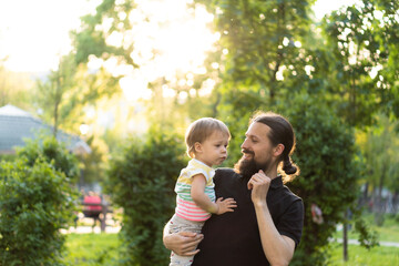 Fatherhood, parenthood, childhood, caring, summer and leisure concept - young dad with beard and long hair in black t-shirt holds in his arms little son in the backlight of the sunset in the park.