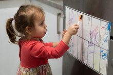 Childhood, Infants Concept - Little Blonde Minor Preschool Girl 2-3 Years Old In Dress Draws With Marker On Planning Board On Refrigerator In Kitchen. Baby Toddler Get Creative On Fridge With Magnets