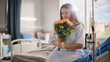 © Gorodenkoff - Hospital Ward: Happy Smiling Latin Female Patient is Sitting on Bed and Holding Received Yellow Flowers with Good Analysis Results. Her Treatment is Already Done. Woman Recovering after Surgery.