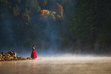 Woman Standing At Lakeshore In Forest During Foggy Weather
