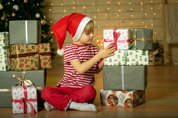 Smiling child in a red Santa Claus hat prepares a Christmas present on the background of a Christmas tree. New Year's magic. Christmas concept