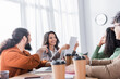 © LIGHTFIELD STUDIOS - Hispanic office worker pointing with finger at document in hands of smiling co-worker in office on blurred foreground