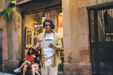 Wall Mural - Cheerful curly hipster guy in casual wear walking on city street checking notification on touchpad enjoying free time, smiling handsome man blogger sending text messages and chatting outdoors