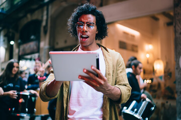 Wall Mural - Expressive handsome male blogger in eyewear stressed with getting message on tablet connected to 4G, emotional man 20s stressed with news from social media on touchpad outdoors on neon light