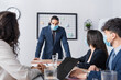 © LIGHTFIELD STUDIOS - Hispanic businessman in medical mask looking at colleagues during meeting in office