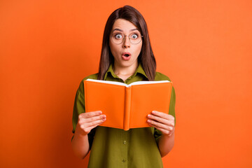 Photo portrait of amazed astonished brunette girl wearing eyewear holding book opened mouth isolated on bright orange color background