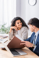 Wall Mural - Hispanic businesswoman smiling at colleague with papers near laptop on blurred foreground