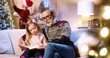 © VAKSMANV - Happy old grandfather sitting on sofa with little cute girl and reading book to his granddaughter at home while his son and grandson decorating xmas tree on background. Happy holidays concept