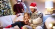 © VAKSMANV - Portrait of joyful caring Caucasian dad with little girl sitting in cozy room at home and reading book. Mom and son decorating glowing Christmas tree. Family holiday time concept. New Year celebration