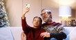 © VAKSMANV - Close up of cheerful smiling Caucasian old grandfather with teen grandson sitting on sofa and taking selfie photos on smartphone at home near glowing cristmas tree. Xmas holidays concept