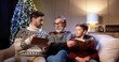 © VAKSMANV - Close up of happy Caucasian family sitting at cozy home and spending Christmas Eve reading book and using tablet. Grandpa talking with son and grandchild having fun. Holiday celebration concept