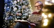© VAKSMANV - Portrait of old nice Caucasian male pensioner grandpa in glasses and christmas sweater reading book while sitting in decorated room near glowing christmas tree. Xmas magic. Winter holidays concept.