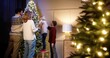 © VAKSMANV - Caucasian happy family with children together decorating xmas tree with twinkle lights putting on decorations in cozy room at home. Mom and dad with kids preparing for Christmas Eve