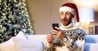 © VAKSMANV - Close up portrait of joyful Caucasian handsome man in santa hat sitting in cozy room near xmas tree typing on smartphone with surprised face win christmas lottery. Happy New Year. Merry Christmas