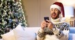 © VAKSMANV - Close up portrait of joyful Caucasian handsome man in santa hat sitting in cozy room near xmas tree typing on smartphone with surprised face win christmas lottery. Happy New Year. Merry Christmas