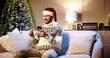 © VAKSMANV - Cheerful Caucasian handsome man in santa hat sitting at home with christmas tree and chatting on smartphone congratulating friends with holidays. Happy New Year. Merry Christmas concept