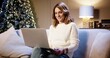 © VAKSMANV - Portrait of young beautiful cheerful Caucasian woman sitting on sofa at home near glowing christmas tree and talking on video chat online on laptop on Xmas Eve. Happy holidays concept