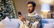 © VAKSMANV - Close up portrait of happy Caucasian young man wearing xmas sweater sitting in room near decorated glowing christmas tree video chatting online on cellphone. Happy New Year. Merry Xmas concept