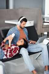 african american woman using phone and lying on bearded man in headphones with laptop in departure lounge