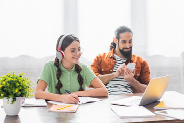 Wall Mural - Smiling hispanic girl looking at laptop and writing in copybook, while sitting near father at table in kitchen