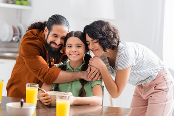 Wall Mural - Hispanic parents hugging smiling daughter during breakfast on blurred foreground