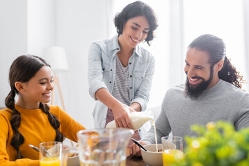 Wall Mural - Smiling hispanic woman pouring milk in cereals near daughter and husband in kitchen