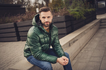 Wall Mural - Photo of good mood attractive young guy dressed green coat having rest sitting stair outdoors city street