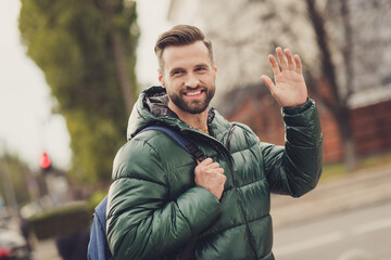 Poster - Photo of cheerful toothy beaming young guy dressed green coat rucksack walking waving arm outdoors urban city street