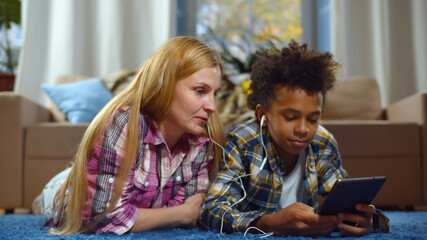 Happy foster mother and son lying on floor and using tablet pc and headphones at home