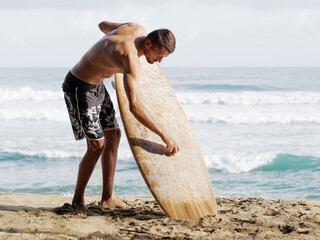 Surfer dressed in boardshorts waxing brown surfboard on the beach at morning. Lifestyle.