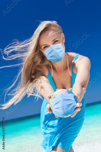 Woman wearing medical mask holding a globe with mask in hands on beach